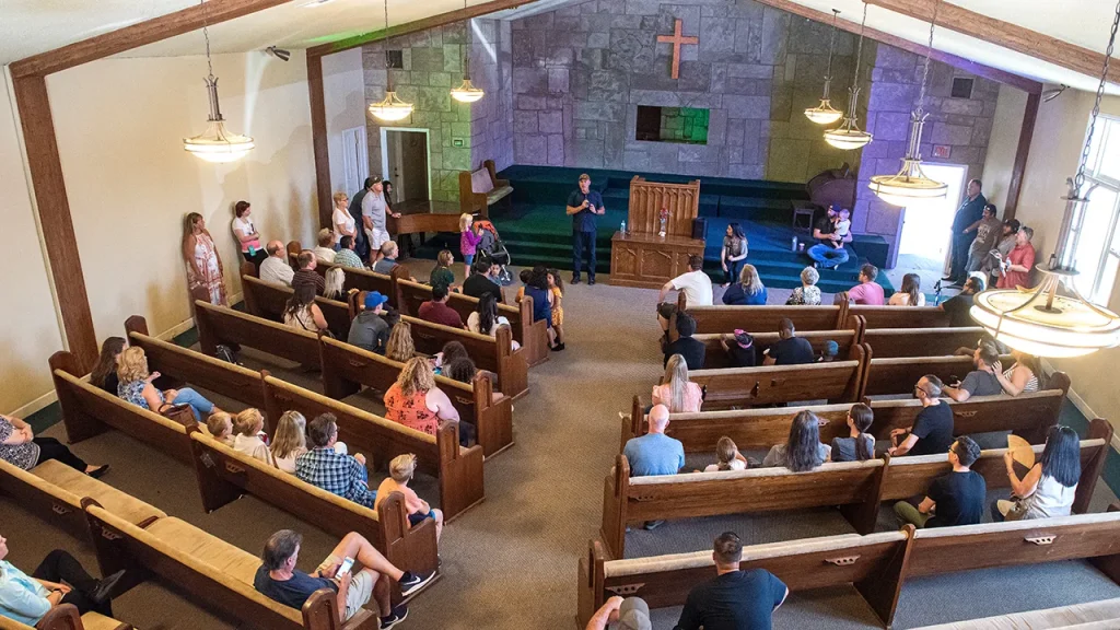 A group of people sits in wooden pews inside a church, watching someone speak at a podium. The church interior features a large cross on the back wall and hanging chandeliers. The congregation includes adults and children, with some standing and some seated.