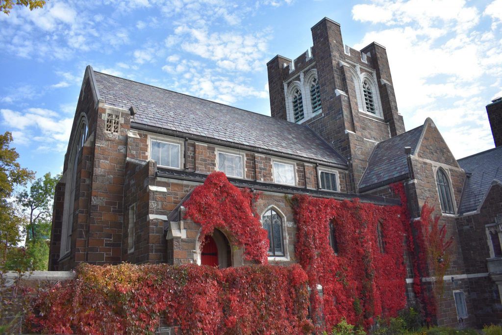 A large, historic stone building with a slate roof is partially covered in vibrant red ivy. The building features arched windows and a tall, square tower. The sky above is blue with scattered clouds. Bushes and trees surround the structure.