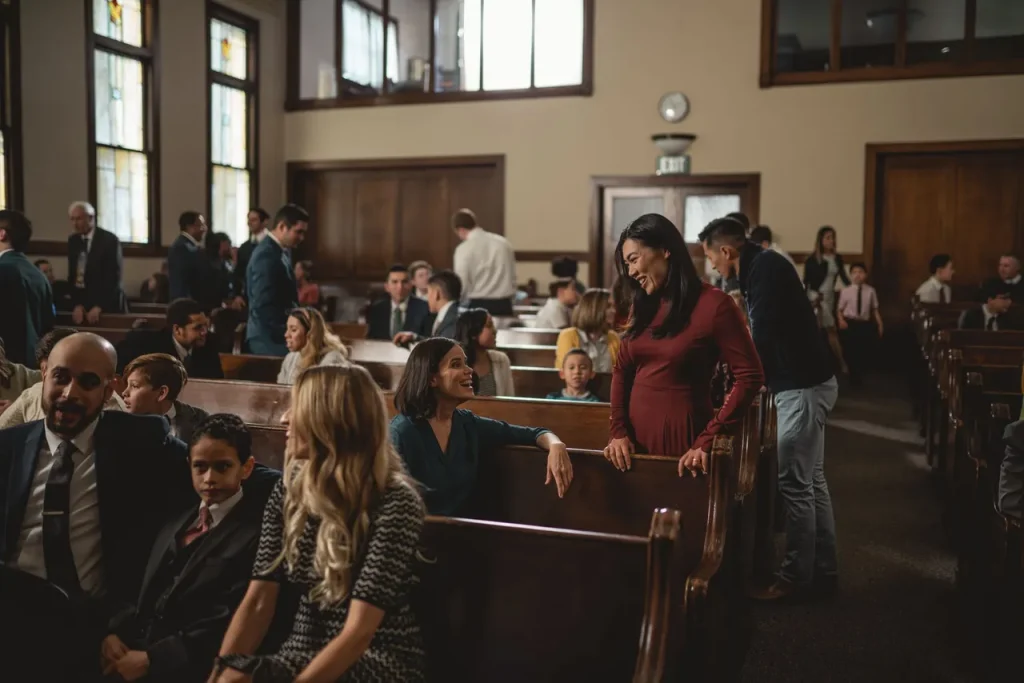 A diverse group of people socializing and conversing in a church or community hall with wooden pews and stained glass windows. Some are seated and chatting, while others stand and engage in discussions. A large clock is visible on a wall in the background.