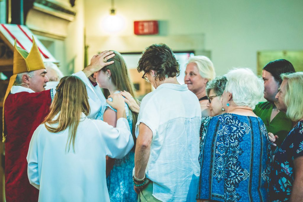 A religious ceremony inside a church where a person in red and gold vestments, likely a bishop, is performing a blessing or confirmation by placing a hand on a young woman's head. A small group of people, including women and elders, stands around them in a supportive gesture.