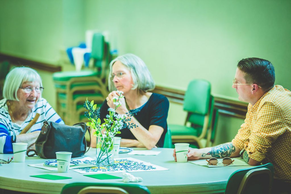 Three people sit around a table engaged in conversation. On the table are some flowers in a vase, disposable cups, and plates. The background shows stacked chairs and a wall. The overall atmosphere appears to be relaxed and informal.