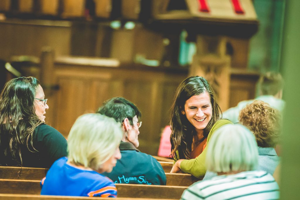 A group of people sits in wooden pews inside a church, engaged in conversation. In focus is a smiling woman with long brown hair, turning to talk to others around her. The background features wooden furniture and church decorations.