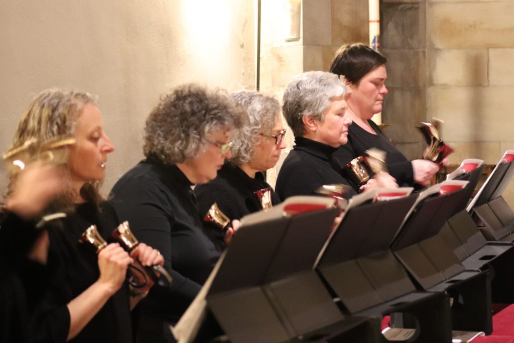 A group of six people wearing black outfits are playing handbells in unison. They stand in a line, each concentrating on their music stands, which hold sheet music. The setting appears to be indoors, with a stone wall in the background.