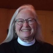 An elderly woman with long white hair, wearing glasses and a black outfit with a white clerical collar, smiles warmly at the camera. The background is softly lit with warm-toned walls.