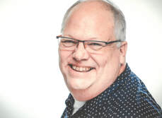 A cheerful man with short gray hair, glasses, and a receding hairline is smiling broadly. He is wearing a dark blue polka-dotted shirt and is posed against a plain white background.