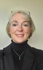 A woman with short blonde hair, wearing glasses on her head, a black turtleneck, and a black blazer is smiling at the camera. She has a silver necklace with a green pendant. The background is plain and light-colored.