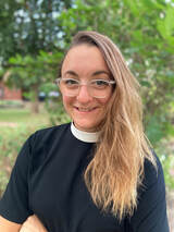 A person with long wavy hair, wearing glasses and a clerical collar, smiles at the camera. The background shows greenery with trees, suggesting an outdoor location.