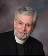 A man with gray hair and a beard is dressed in clerical attire, including a black shirt and white clerical collar. He is smiling slightly and is posed against a neutral, blurred background.