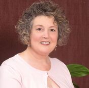 A woman with short, curly gray hair is smiling with a slight tilt of her head. She is wearing a light pink cardigan over a white top, with a dark maroon background behind her and a green plant partially visible to her right.