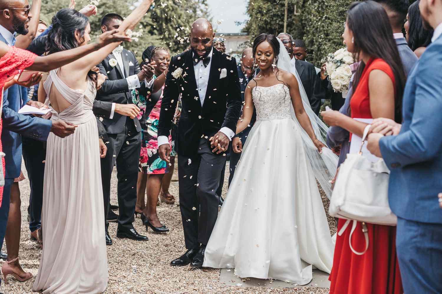 A newlywed couple walks down an outdoor aisle, smiling and holding hands. The bride wears a strapless white gown, and the groom is in a black suit. Guests, dressed in various colors, cheer and throw flower petals. The background features greenery and trees.