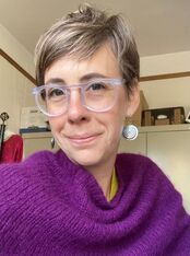 A person with short, light-colored hair and wearing clear-framed glasses smiles at the camera. They are dressed in a purple sweater and large, round earrings. The background shows a room with shelves and a wooden door.