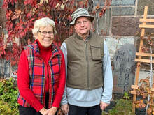 An elderly couple stands together in front of a stone wall covered in red ivy. The woman wears glasses, a red sweater, and a red plaid vest, while the man is in a gray shirt, green vest, and a bucket hat. They smile warmly at the camera.