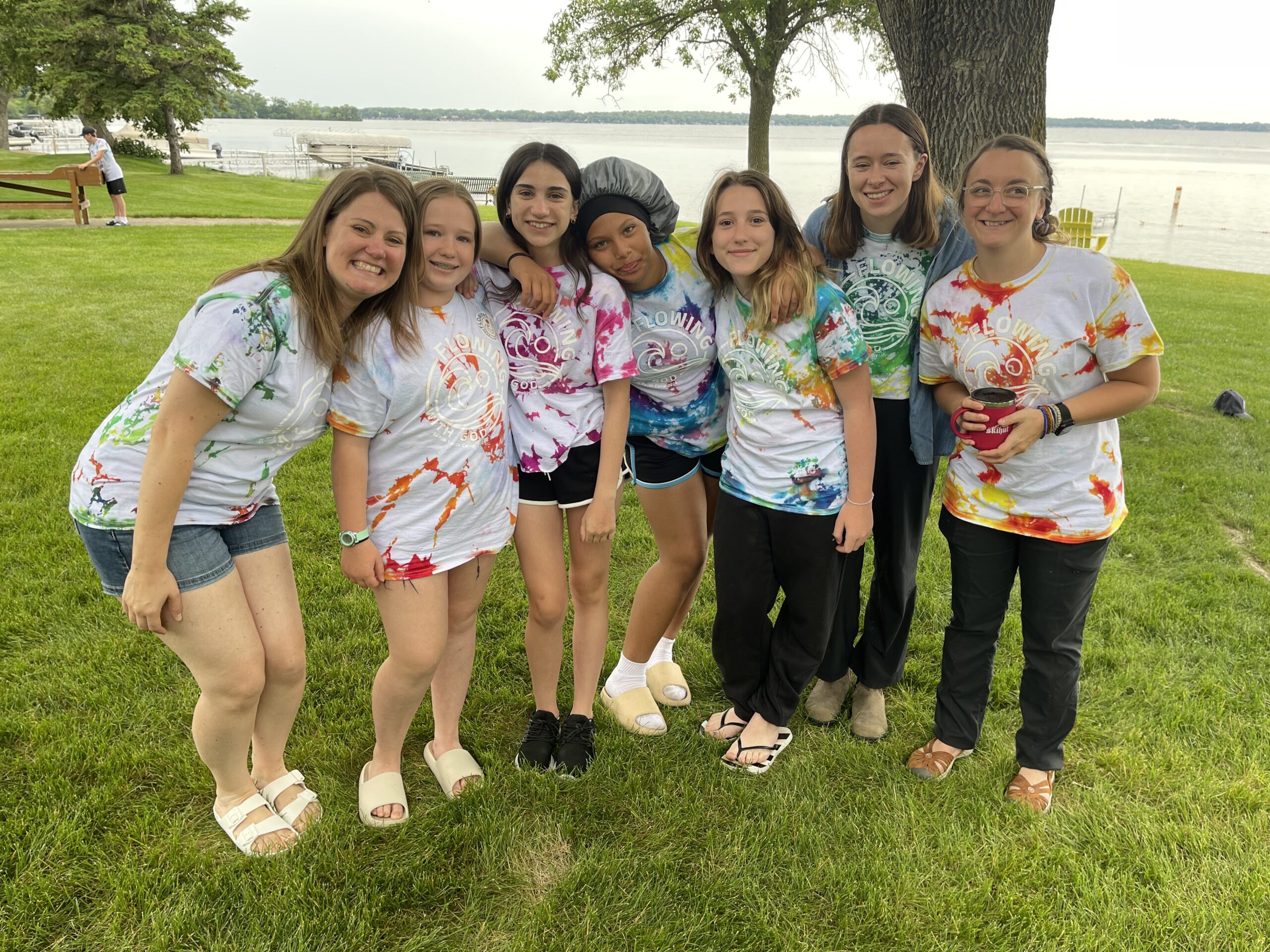 A group of seven people, consisting of adults and children, stands smiling on a grassy area near a body of water. They are wearing colorful tie-dye shirts. Trees and picnic tables are visible in the background.