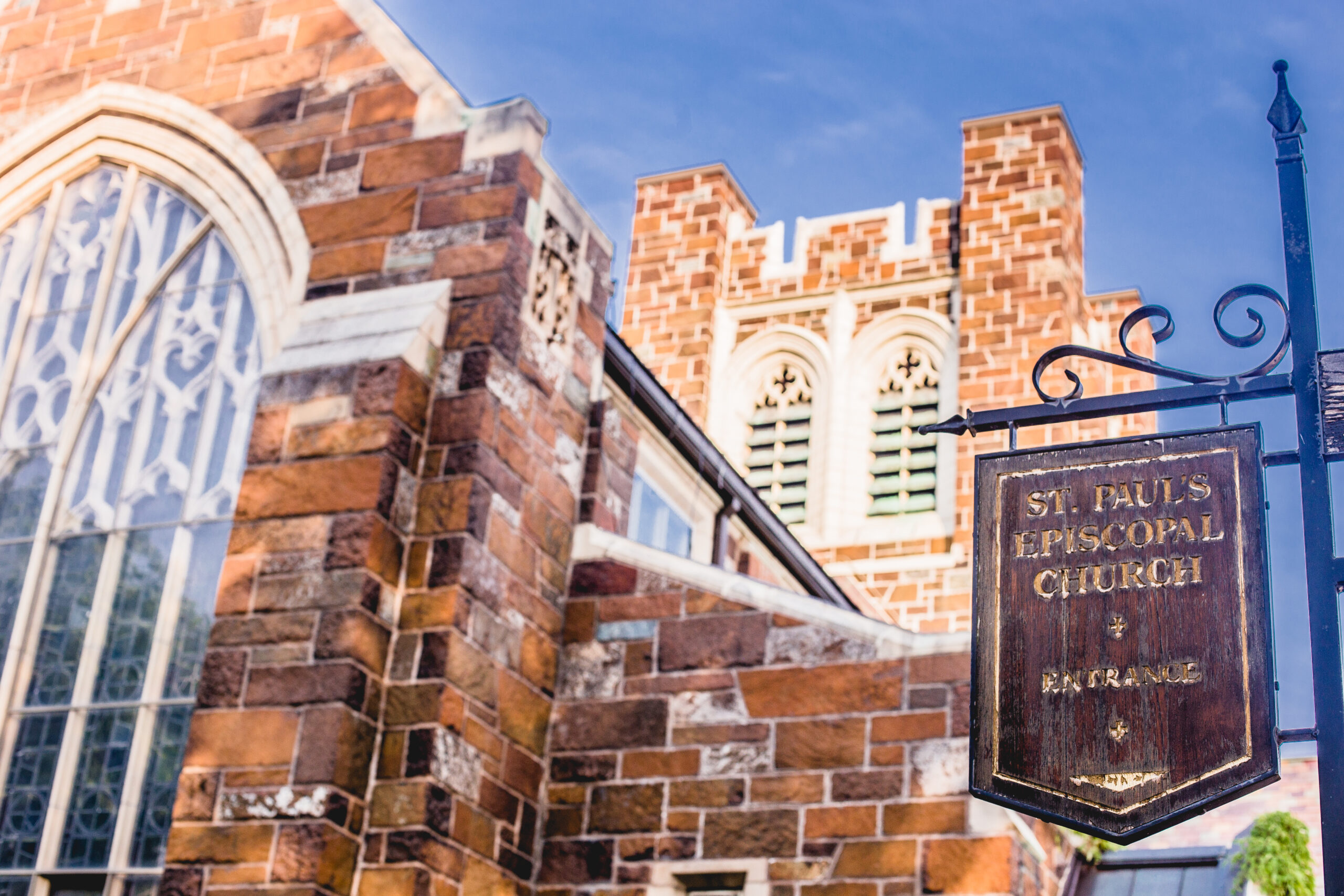 A brick and stone church with a large arched window on the left and a tower with decorative openings in the background. A wooden sign in the foreground reads 