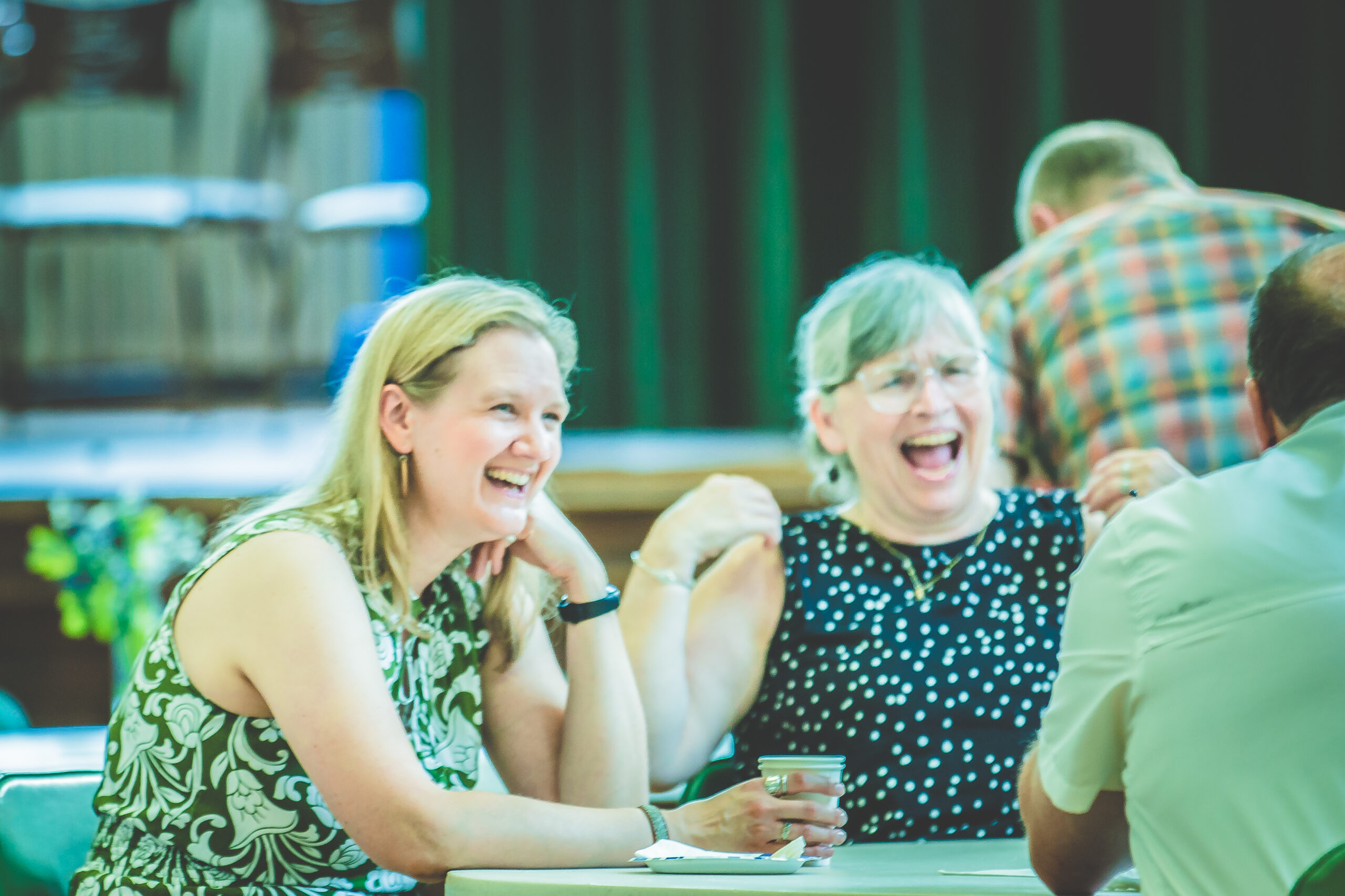 Two women are sitting at a table, laughing heartily, during a social gathering. Both are wearing sleeveless tops; one has a patterned top and is resting her elbow on the table. The background shows a blurred setting with a third person partially visible.