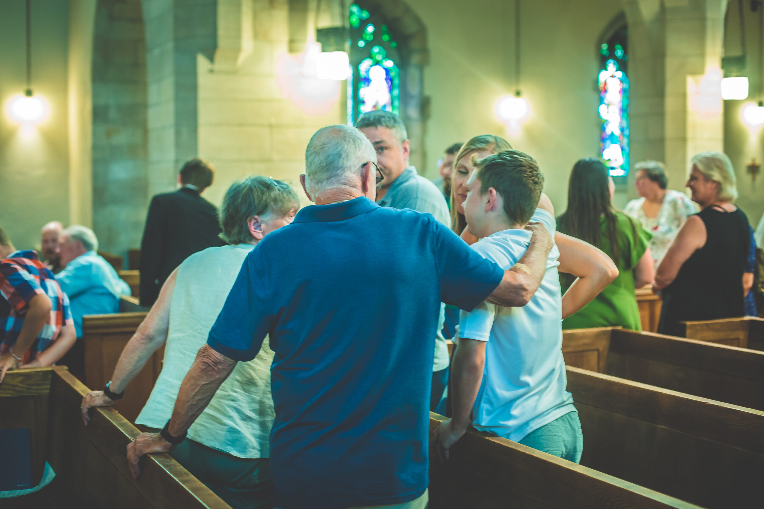 A group of people is gathered inside a church, standing and sitting in wooden pews. An elderly man with a hand on a younger person's shoulder is seen from the back. The background features stained glass windows and soft lighting.