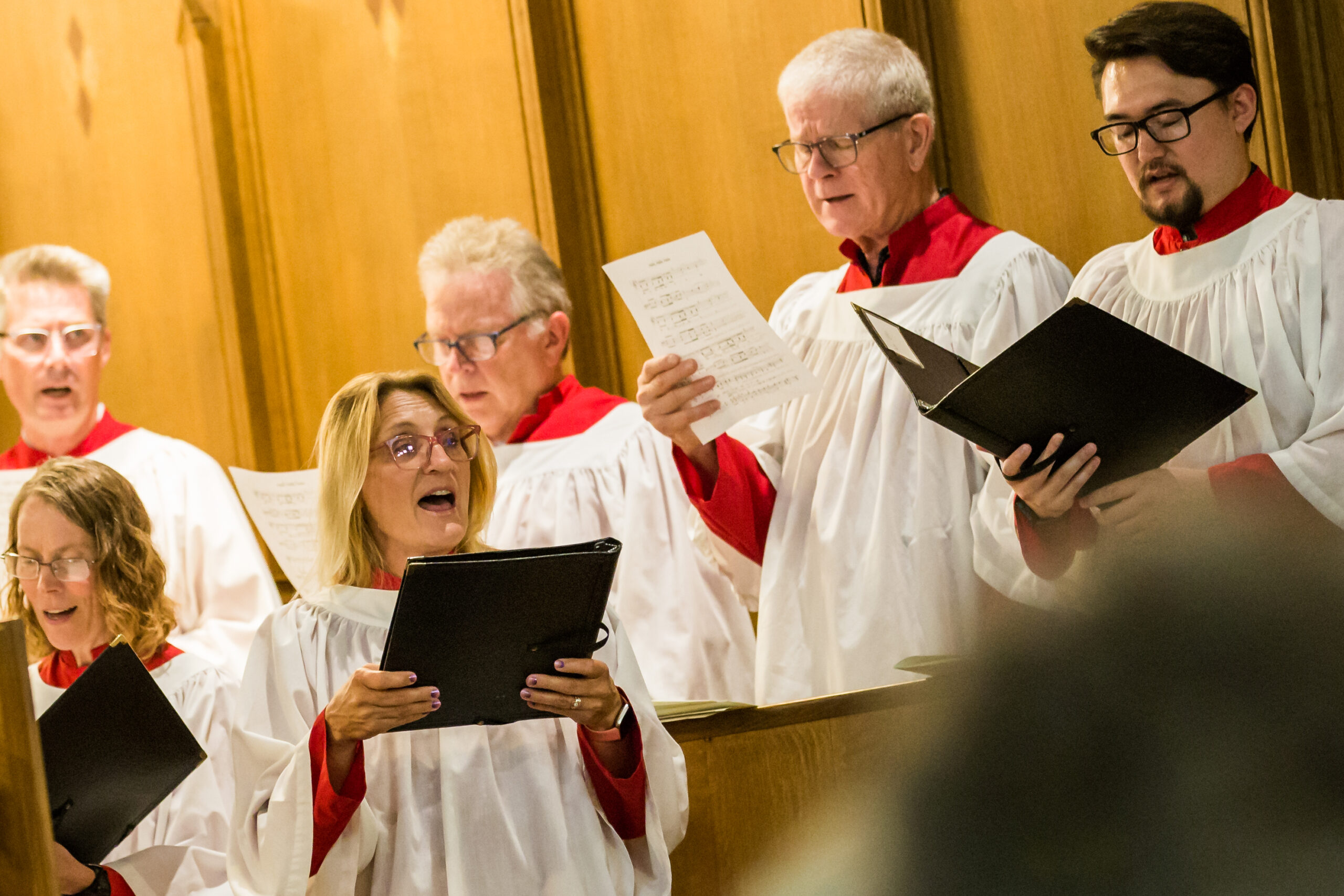 A group of people dressed in white choir robes with red collars sing in a choir. They are holding black folders with sheet music. The choir members are varying in age, and they are standing in front of wooden paneling.