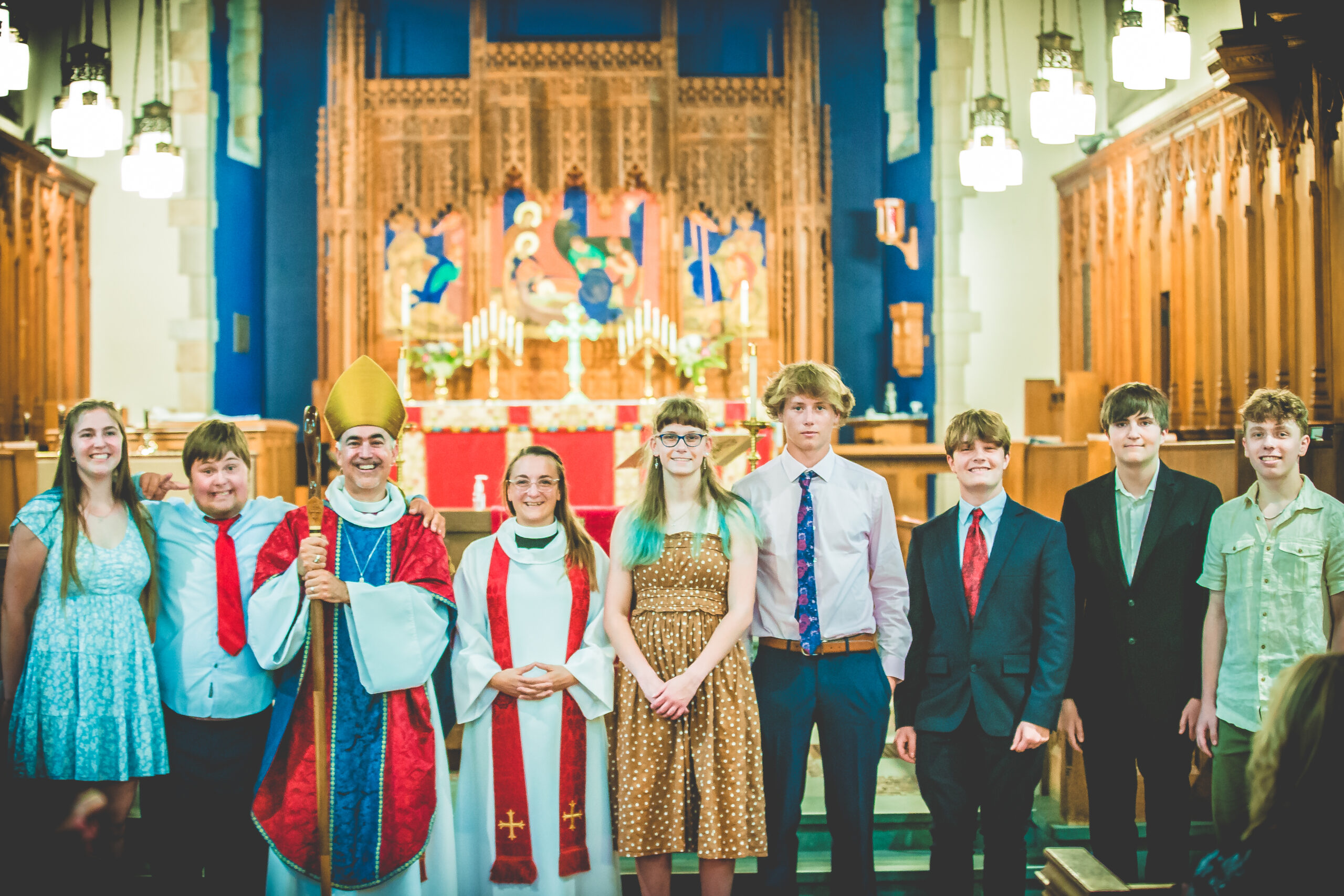 A group of eight people dressed in various formal attires stands inside a church. Two clergy members in religious garments, including one holding a staff and wearing a mitre, are in the middle. The background showcases an ornate altar with candles and religious decorations.