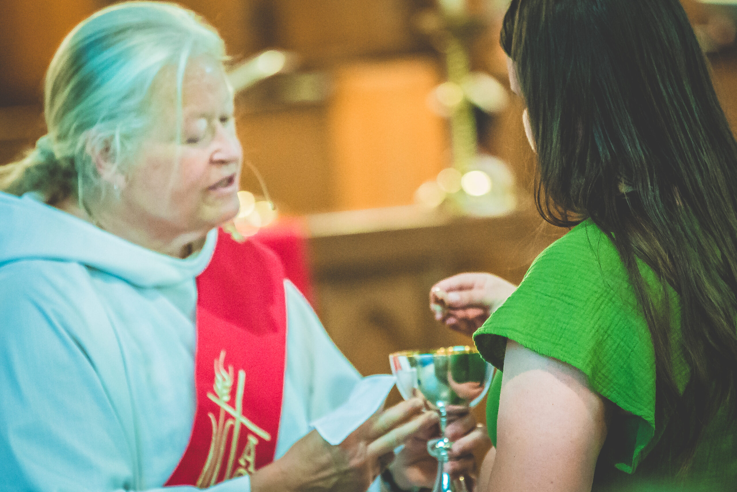 A person in a religious white robe and red stole offers communion to a participant in a green dress inside a church. The participant is taking a wafer from the priest, who is holding a chalice. The background is blurred with warm lighting.