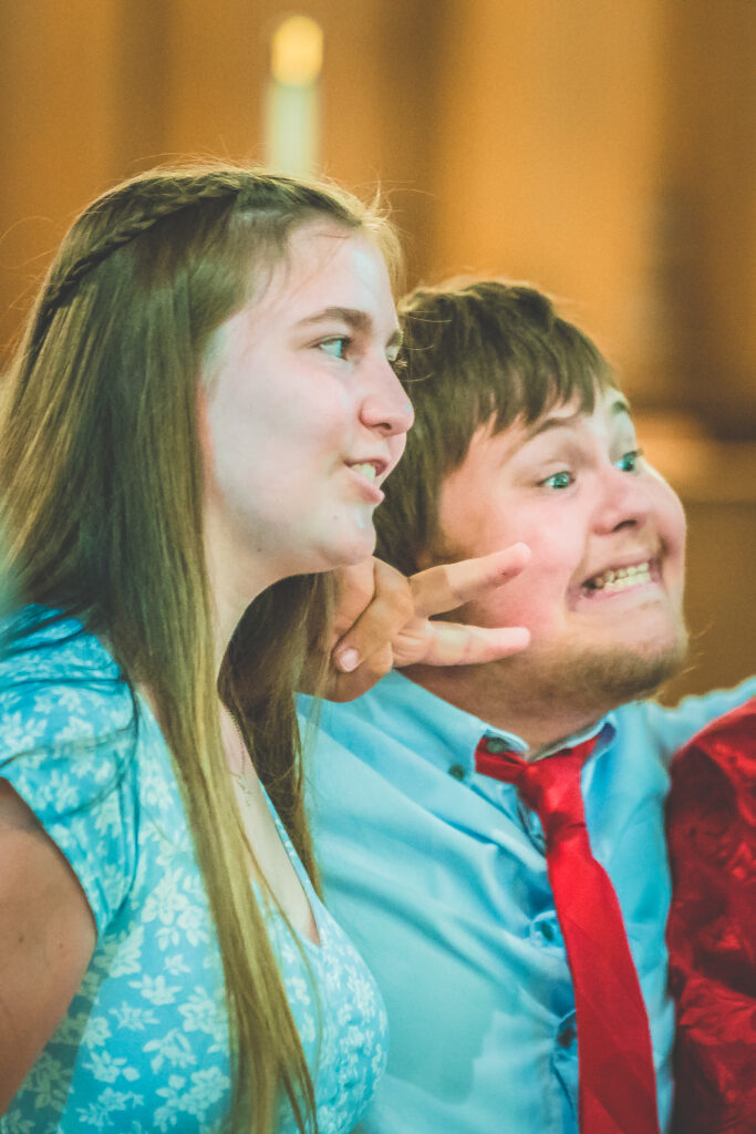 Two smiling young people are posing for a photo. The girl on the left has long straight hair and is wearing a light blue patterned dress. The boy on the right, in a light blue shirt and red tie, is making a peace sign with his hand on her shoulder. They appear happy and excited.