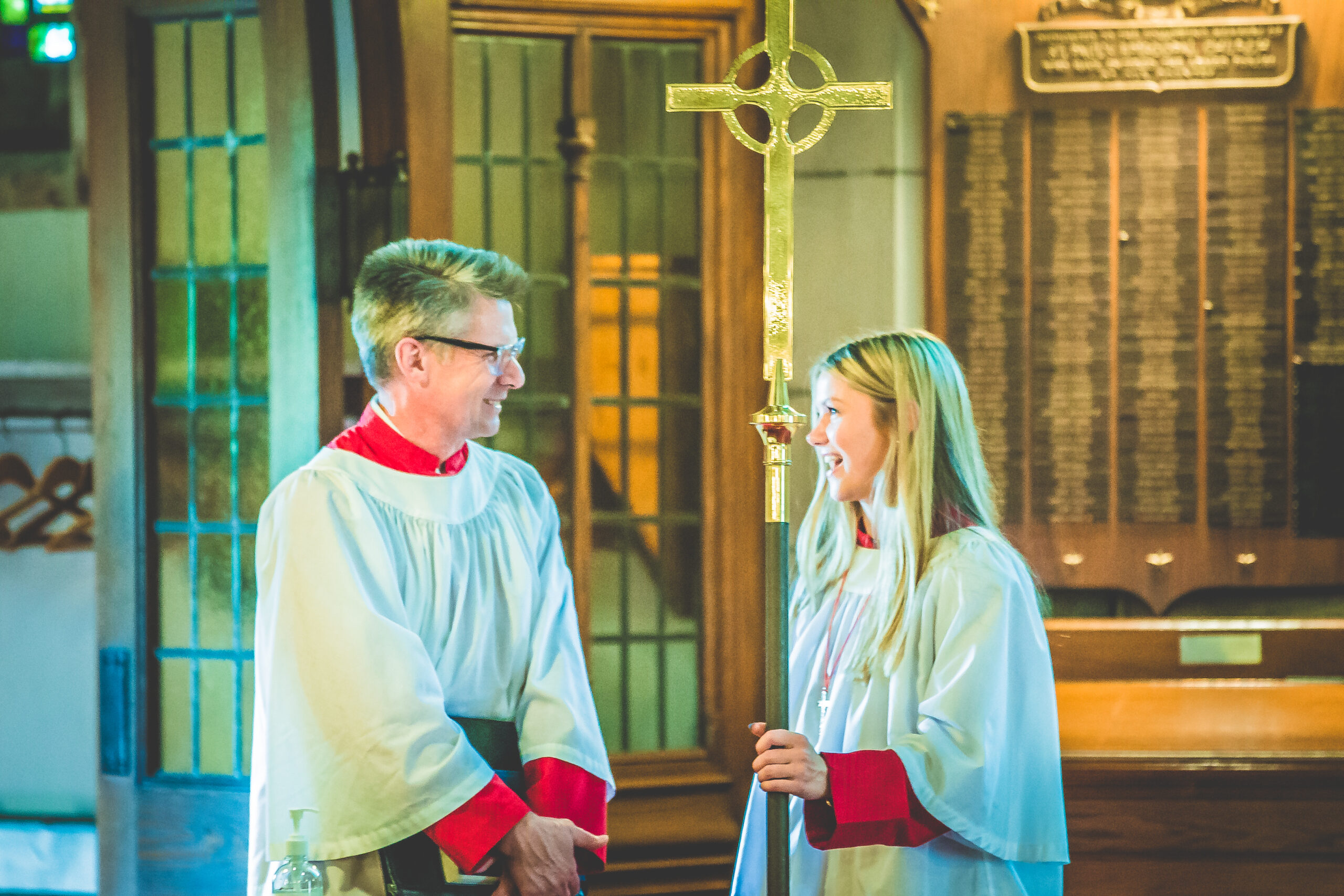 Two people dressed in white and red liturgical robes stand inside a church. One person, on the right, holds a large gold cross staff. They are smiling and looking at each other, bathed in soft, warm light. Ornate wooden panels and a door are visible in the background.