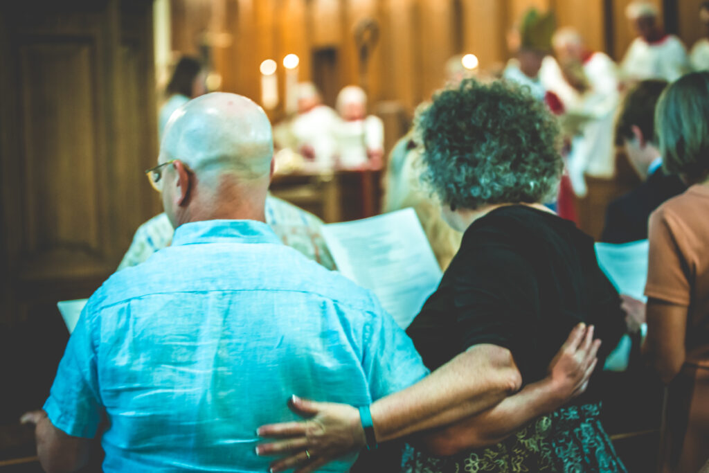 A group of people standing closely together in a church, holding papers and singing or reading. Some are embracing each other. Focus is on the back of two individuals in the foreground, with an altar and clergy visible out of focus in the background.