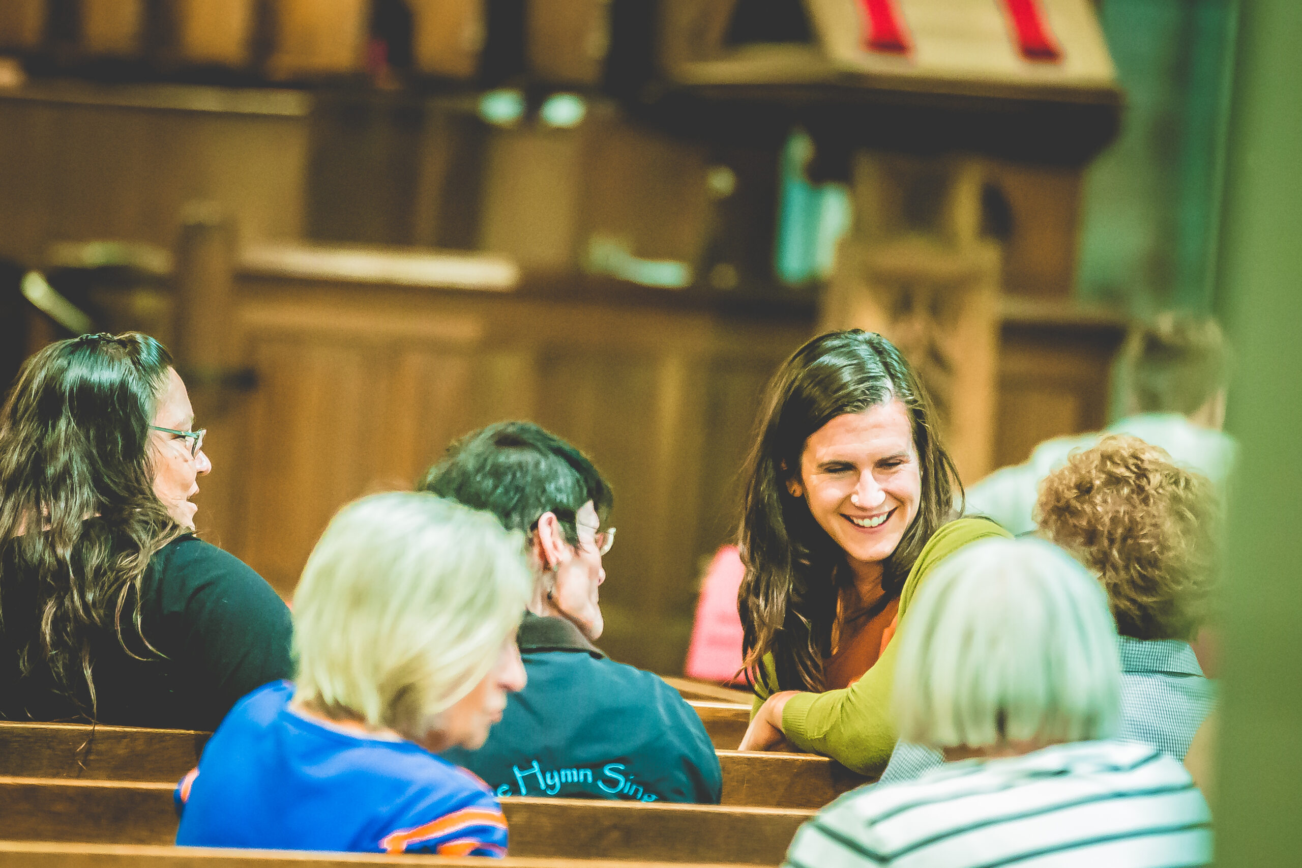 A group of people sitting in wooden church pews, engaged in conversation. A smiling woman with long brown hair, wearing a green top, sits in the middle of the group. The background shows the church's wooden interior and part of an altar.