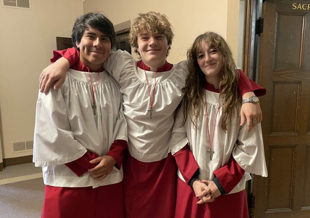 Three young people standing in a hallway, each wearing red robes with white overgarments and silver cross necklaces. They are smiling, with the person in the center having their arms around the shoulders of the other two. A wooden door labeled 