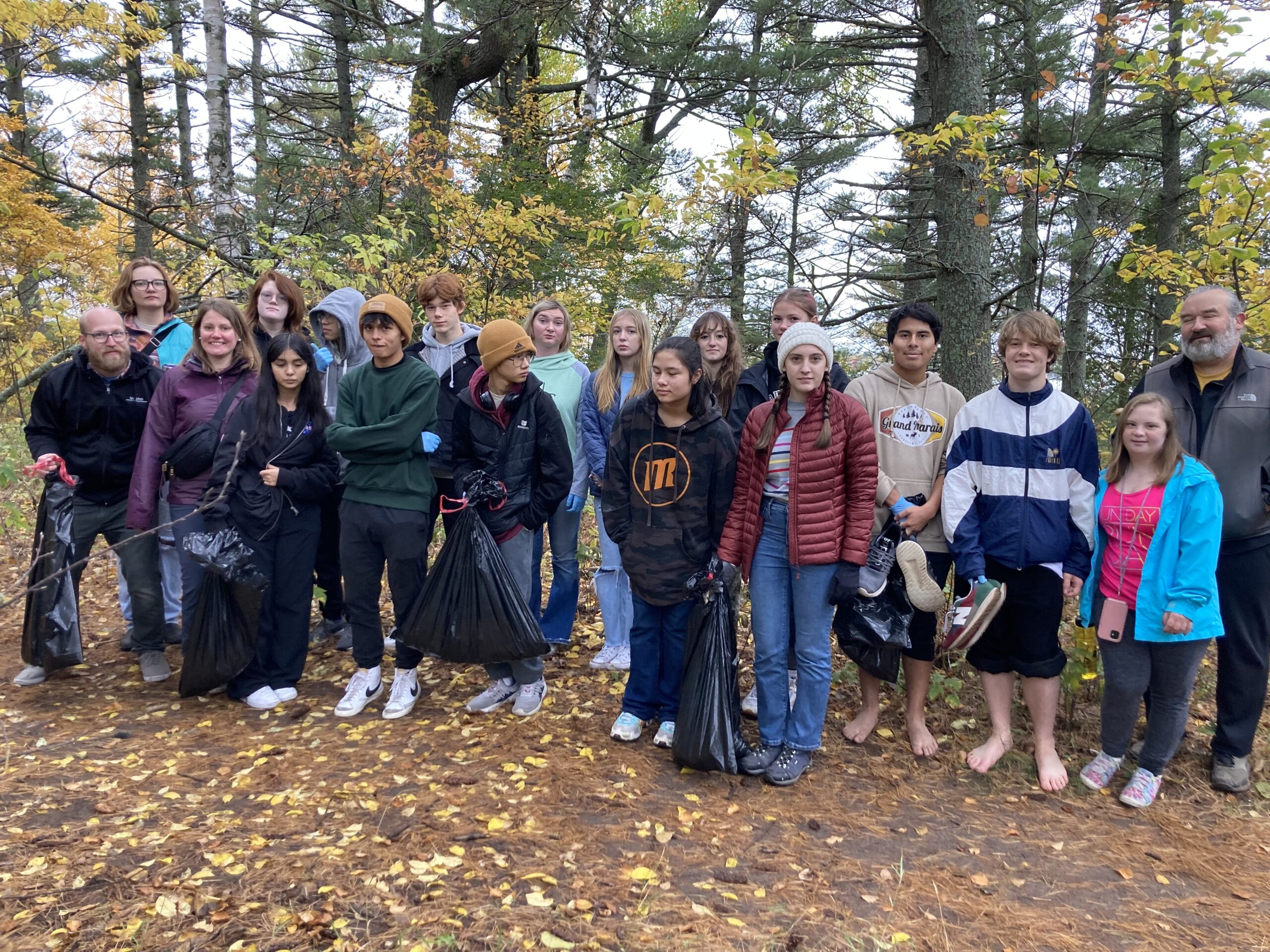 A group of people stands outdoors in a forested area during fall. They are holding black trash bags, suggesting they are participating in a cleanup activity. The group consists of adults and children dressed in casual, warm clothing.