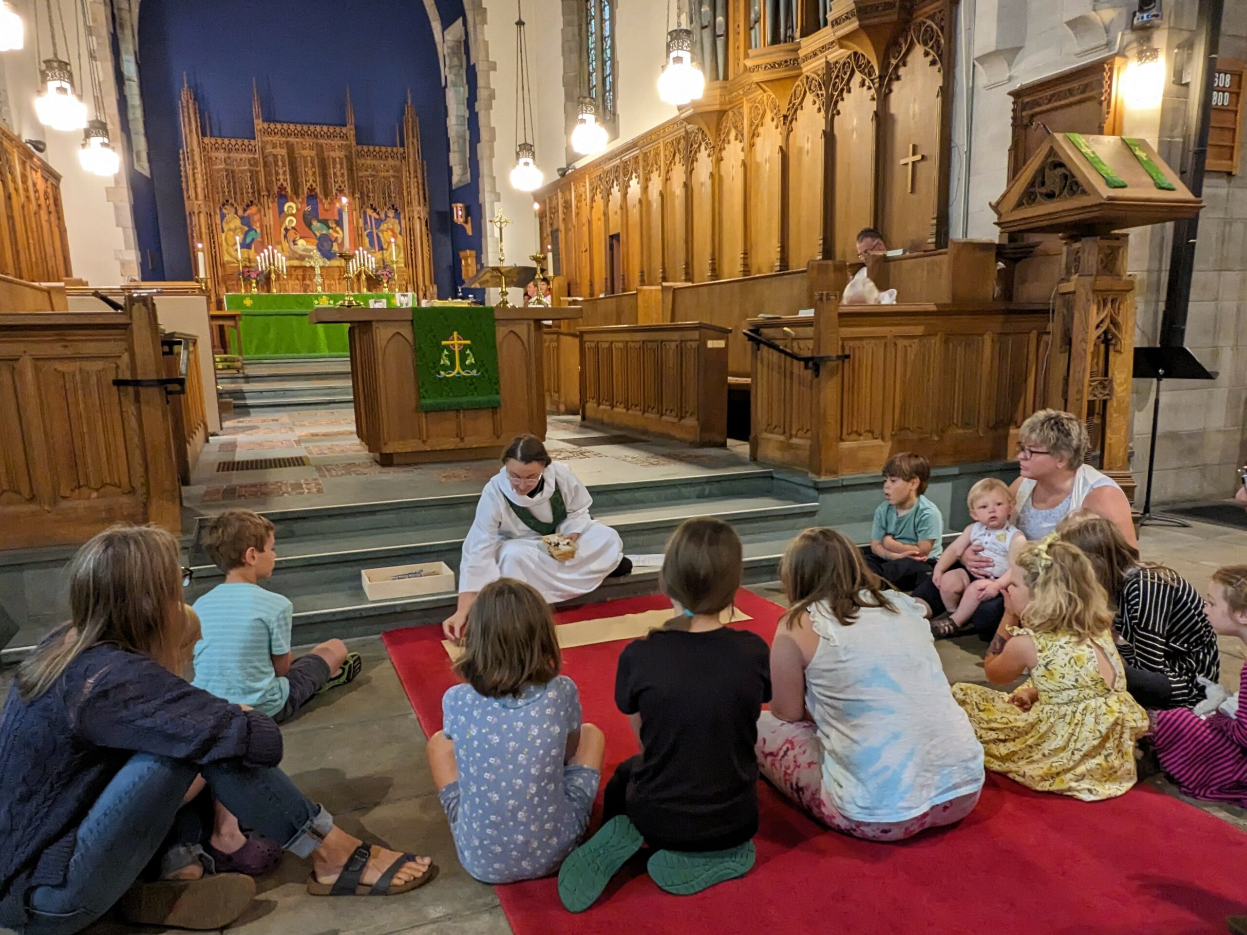 A person wearing religious attire sits on the floor of a church with a group of children and a few adults sitting around them. The group is gathered near the altar, which is decorated with green cloth and lit candles. The scene takes place inside a wooden-paneled church.