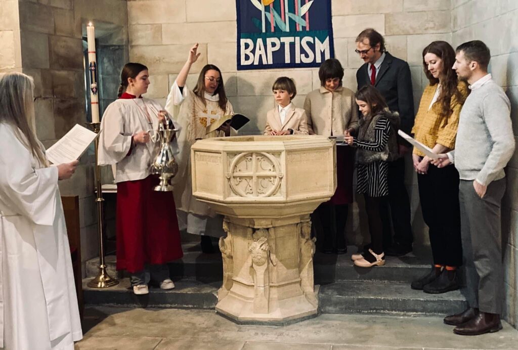 A group of people stand around a stone baptismal font in a church during a baptism ceremony. Two ministers, dressed in white robes, lead the service. Seven other people, including children and adults, are gathered around, with one minister holding a censer.