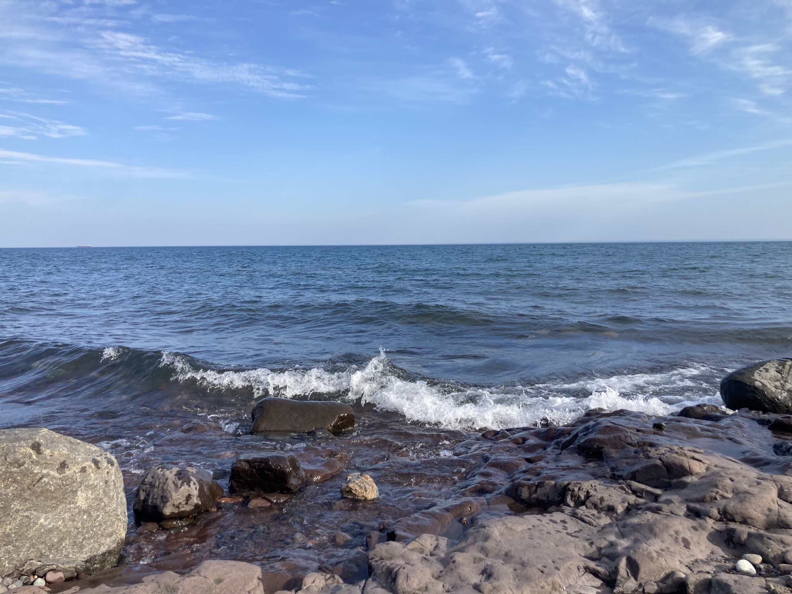 A serene view of a rocky shoreline with waves gently crashing against the rocks. The blue sky above is scattered with light clouds, and the vast ocean extends to the horizon, blending seamlessly with the sky.