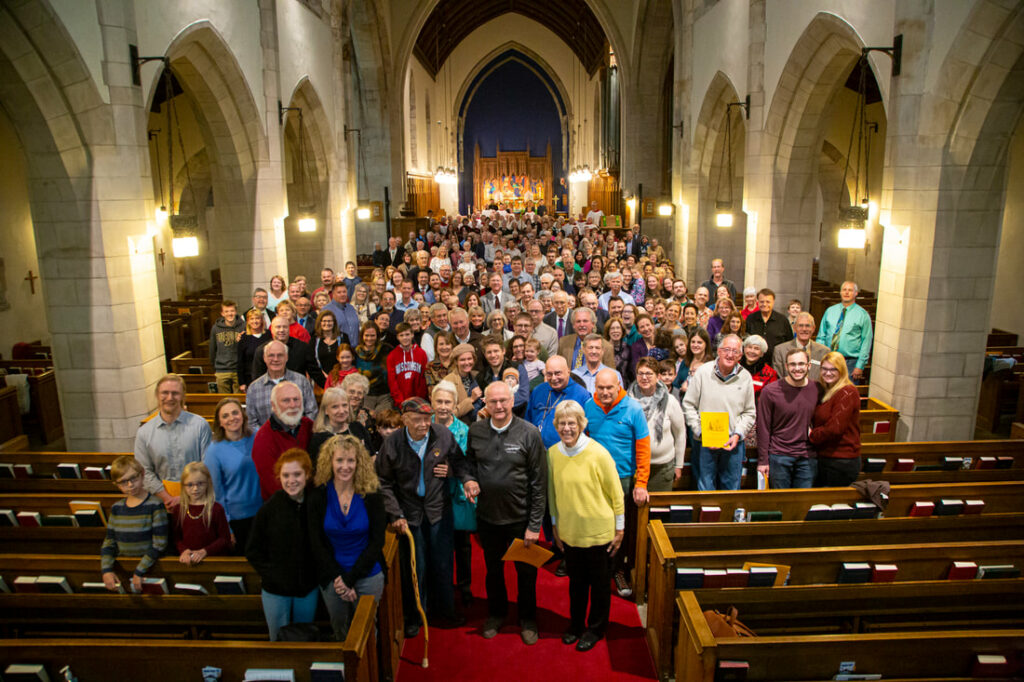 A large group of people gather in the pews of a grand church with vaulted ceilings and arched windows. The crowd spans various ages, smiling warmly towards the camera. The church is warmly lit, enhancing the welcoming atmosphere.