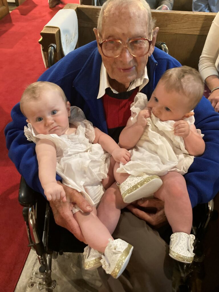 An elderly man with glasses and a blue jacket sits in a wheelchair, holding two babies dressed in white outfits. The babies have light hair and are both looking in different directions. The setting appears to be indoors, possibly a church, with a red carpet visible.