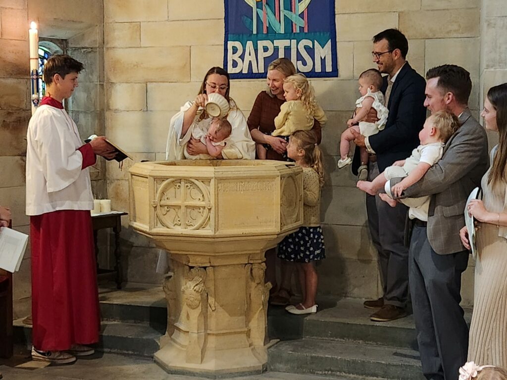 A baby is being baptized with holy water in a church. The officiant pours water from a vessel onto the baby's head over a baptismal font, while people (including parents and siblings) stand close by. A religious banner reading 