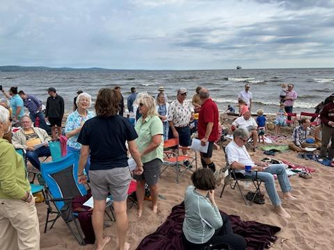 A group of people are gathered on a sandy beach near the ocean. Some are standing and talking, while others sit on colorful folding chairs. The sky is cloudy, and the waves are visible in the background. The atmosphere appears to be casual and relaxed.