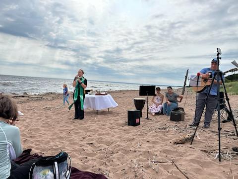 A small group of people gathered on a sandy beach with the ocean in the background, engaged in a casual performance. One person is speaking into a microphone, and another is playing a guitar. There is a table with a white cover and some equipment set up nearby.