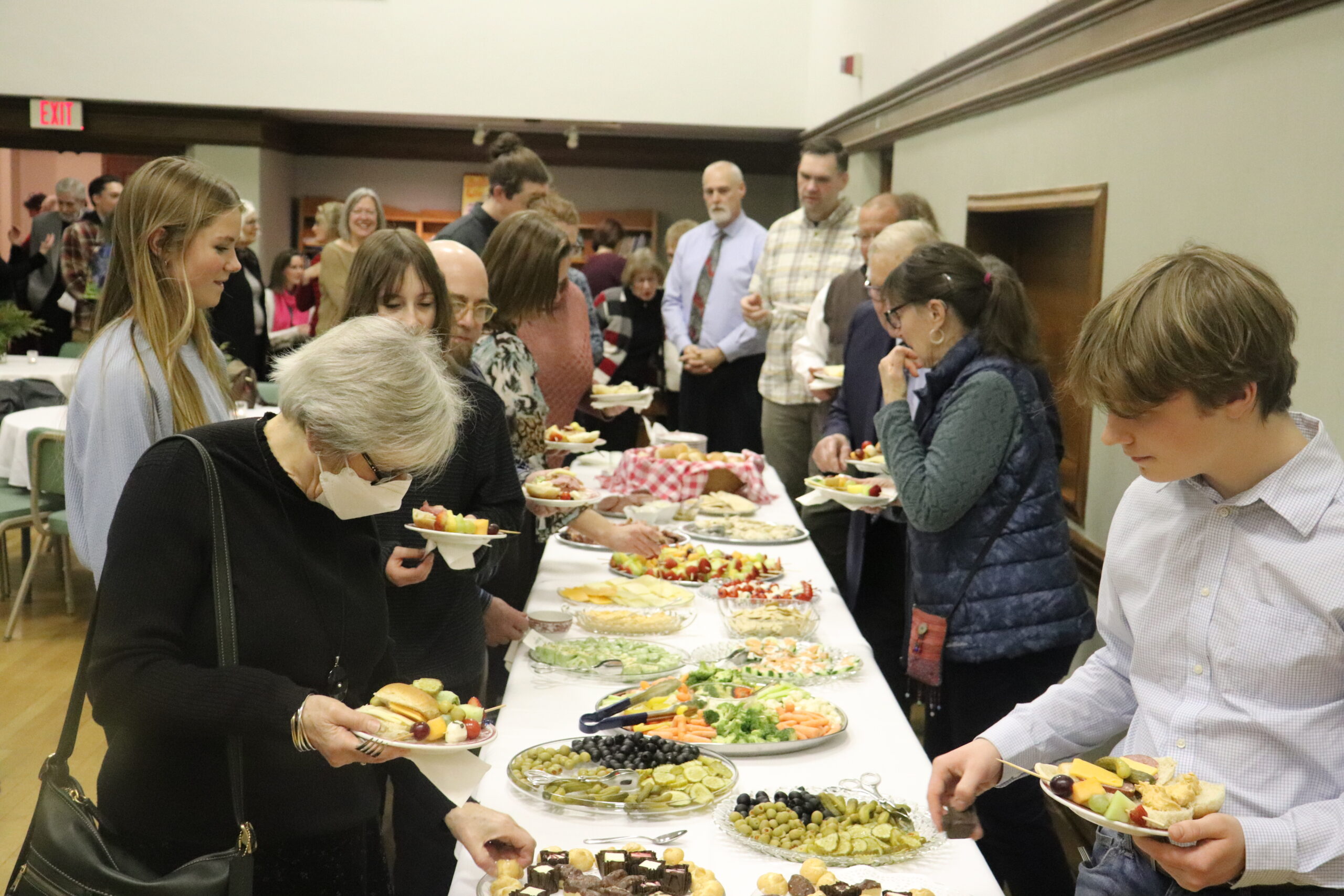 A group of people is gathered around a long buffet table, selecting from a variety of foods. The table is laden with fruits, vegetables, and desserts. People of different ages are present, engaging in conversation as they serve themselves. The setting appears casual and social.