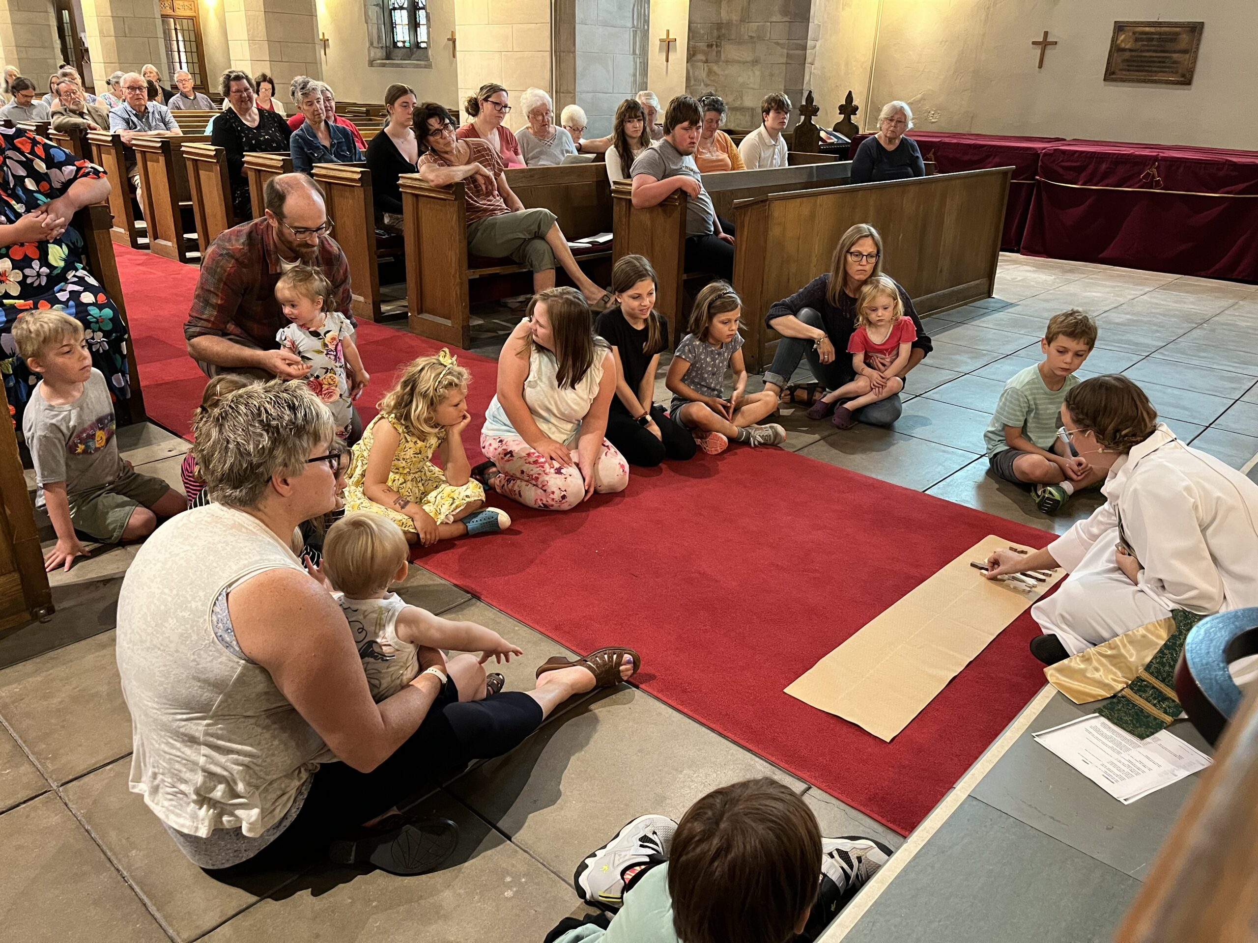 A group of children and adults sit on the floor in a circle inside a church, engaged in an activity led by a person in religious attire. The church pews are occupied by additional attendees observing the scene.