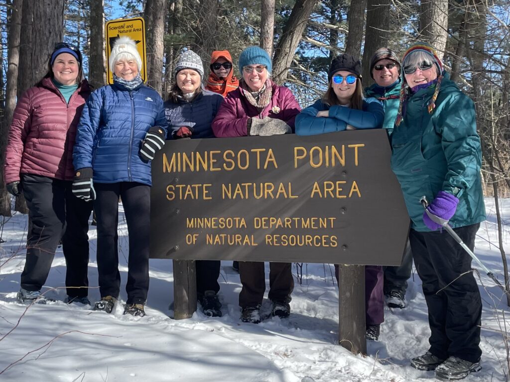 A group of eight people, dressed in winter clothing, stand together smiling in front of a sign that reads 