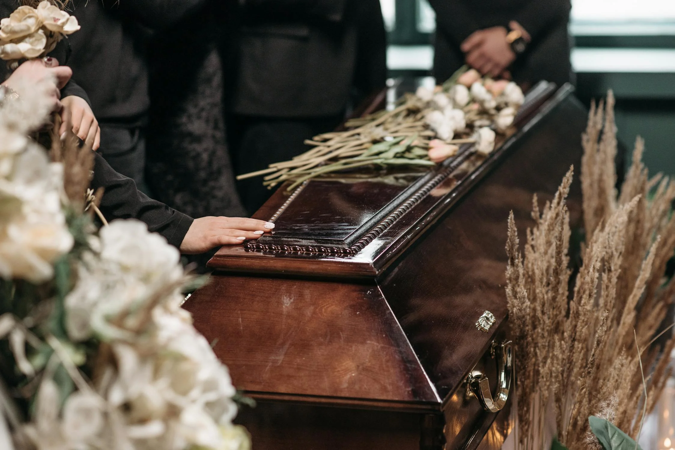 A group of people, dressed in black, stands around a wooden coffin adorned with flowers during a funeral service. One person rests their hand gently on the coffin, surrounded by floral arrangements and muted decor.